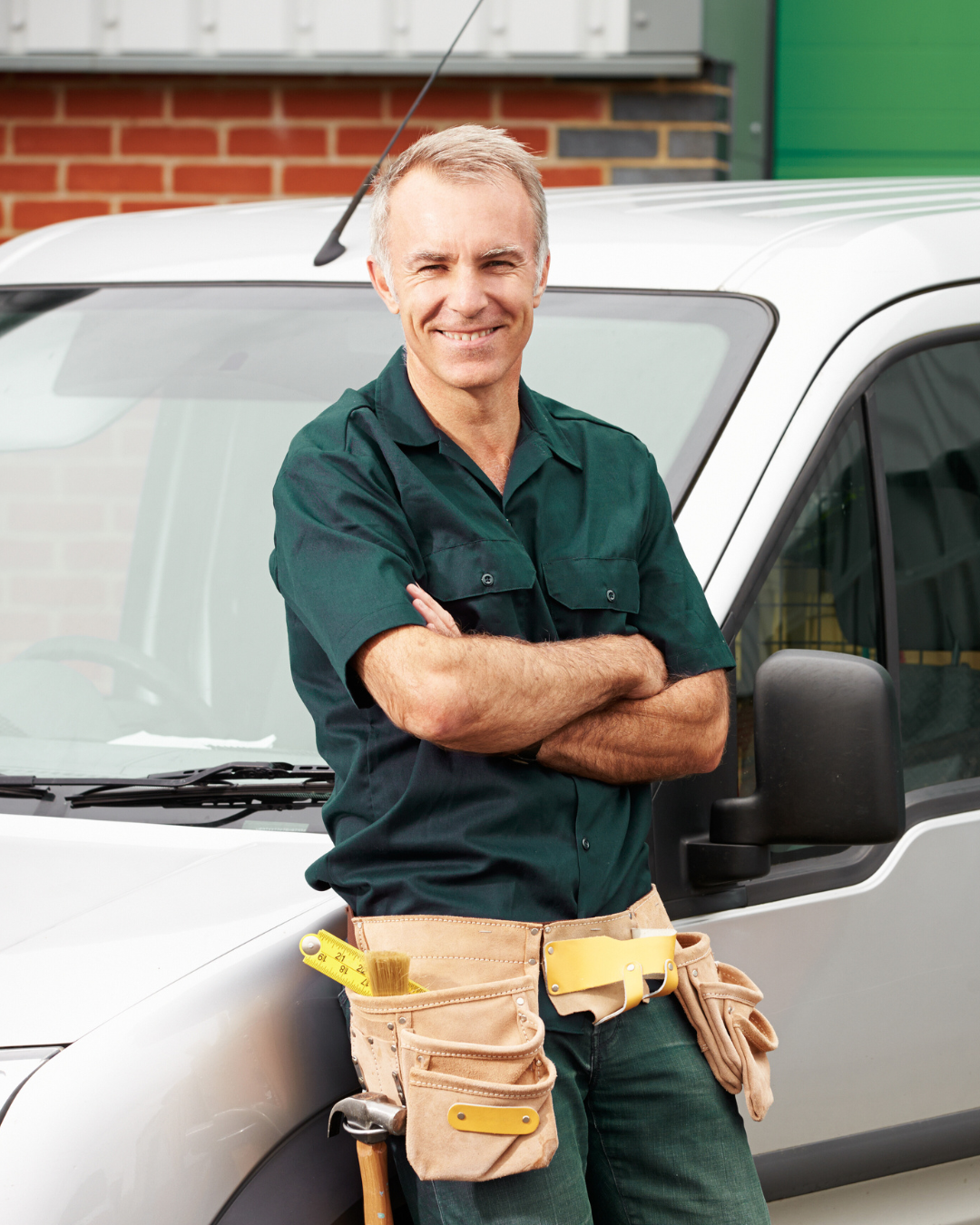 Manual labourer standing next to a white van, smiling, with tools in a tool belt, in front of a brick building.